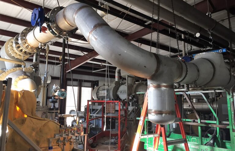 Industrial piping installation with large process lines, valves, supports, ladders, and lift equipment for gas processing system construction.
